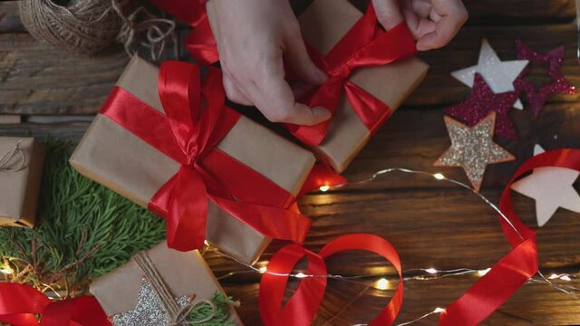 female woman hands wrapping Christmas presents gift box with brown craft paper, tying red ribbon bow on wooden desk with new year ornaments: stars light garland and green coniferous. top view DIY home