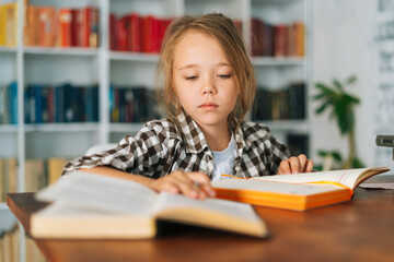 Front view of cute focused pupil child school girl doing homework writing in notebook sitting at table with paper book. Portrait elementary kid schoolgirl studying at home, selective focus..