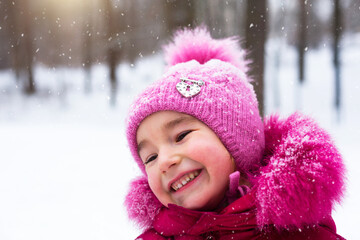 Little girl in the cold looks at the snow and smiles. Winter, walk the baby in the open air, snow. Warm clothing, knitted hat, fur pompom and hood. Snowball fight, winter fun,. Close-up portrait