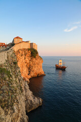 Fototapeta na wymiar Fortress walls of Dubrovnik with a pirate galleon ship sailing in the harbor at sunset