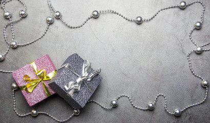 Two shiny gifts of pink and silver color with ribbon bows on a gray concrete background around a string of silver beads with copy space.