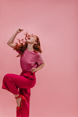 Young woman very energetically rejoices in victory waving hands clenched fists in enclosed space. Fair-skinned red-haired lady bends leg to knee, wearing pink T-shirt, crimson pants and red sandals.