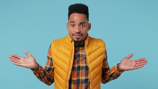 Fun confused shy young african american†man 20s wears yellow shirt waistcoat look around spread hands say oops ouch oh omg i am so sorry isolated on plain pastel light blue background studio portrait