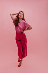 Full length fashionable caucasian young lady posing walking forward holding her waist and hair from behind in pink room. Tipping her head red-haired girl with open mouth in T-shirt, pants, shoes.