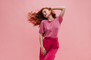 Young girl of bright appearance with red hair flying to side indoors with place for text. Fair-skinned teenager with red lips in light pink T-shirt tucked into crimson pants. Elegant style concept