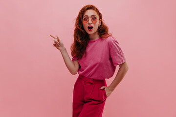 Attractive fair-skinned girl with surprise points her index finger to side on pink background. Stylish redhead woman holds hand in pocket dressed in casual pink clothes. Concept indicating copy space.