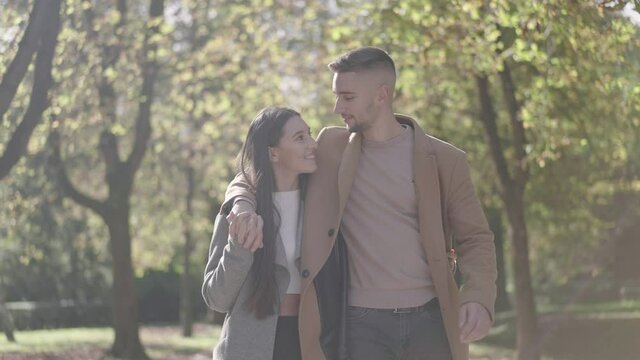 Slow motion - Young man hugging his girlfriend over her shoulder, holding hands, and walking down a path surrounded by tree avenue, on a sunny day