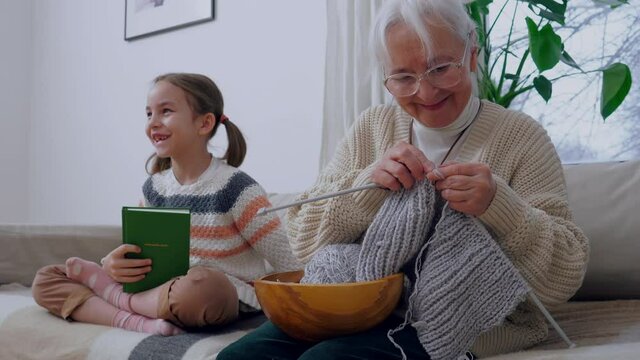 Proud granny sitting on a couch with her granddaughter. They talk while grandma knits a hat