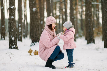 Fototapeta na wymiar Mom with her cute little daughter during winter walk in snowy forest. Mom and child in pink jackets looking each other with love and tenderness