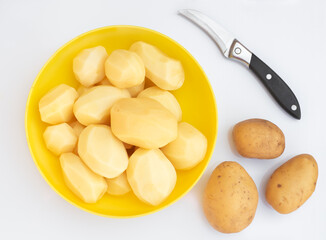 Peeled potatoes in a yellow cup on a light surface. Nearby there is a knife and potato tubers. Cooking