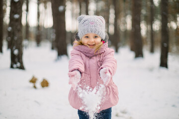 Cute laughing child girl having fun in winter forest. Little girl in pink coat throwing the snow and smiling. Seasonal outdoor portrait of pretty small girl in winter forest