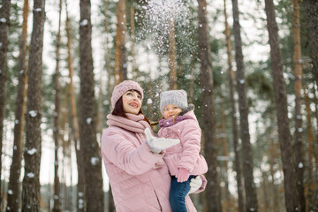 Fototapeta na wymiar Young mother holding cute little daughter, in a winter forest for a walk. Pretty mom with child on hands, wearing pink coats throwing snow up and laughing. Family in winter forest