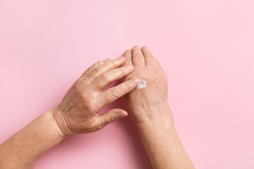 Elderly woman applying cosmetic cream onto her hands against color background