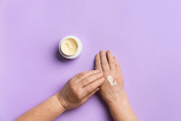 Elderly woman applying cosmetic cream onto her hands against color background
