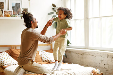 Happy african american father and son jumping on bed and laughing while playing together at home - obrazy, fototapety, plakaty