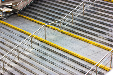 A wide, modern stone staircase with yellow markings for blind people and metal handrails for a...