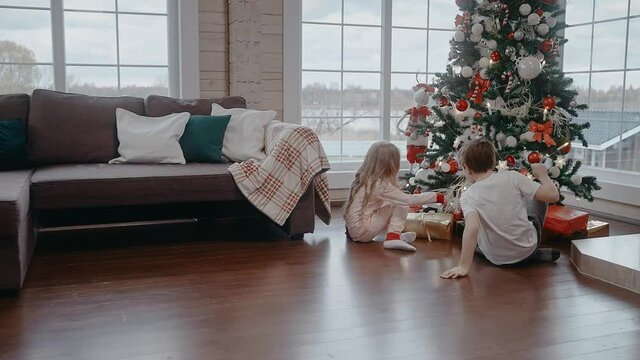 Boy and girl are running to open their gifts under the tree on Christmas morning. Christmas morning in big and bright house. Kids are excited to get their presents. 50 fps