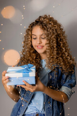 beautiful young woman with blue eyes and blonde curly hair in denim clothes holds a gift box in her hands. girl opens the gift. solid gray background. space for text. High quality photo