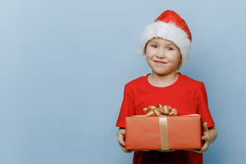 Funny boy in a Santa suit and hat is holding a red gift box in his hands and smiling sweetly, looking at the camera, isolated on a blue background. Merry Christmas gift sale.