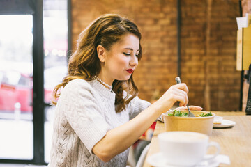 Beautiful young woman eating a salad from a recycling paper bowl while having lunch in the cafe. Healthy habits during the day. Diet planning