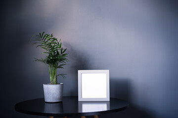 Empty light grey frame, small Areca Palm home plant in a concrete pot on black table with dark gray wall background. Mockup. Modern minimalist interior design. Copy space, selective focus