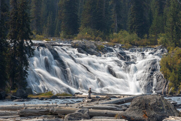Fly fisherman fishing in a river at the base of a large waterfall in the mountains.