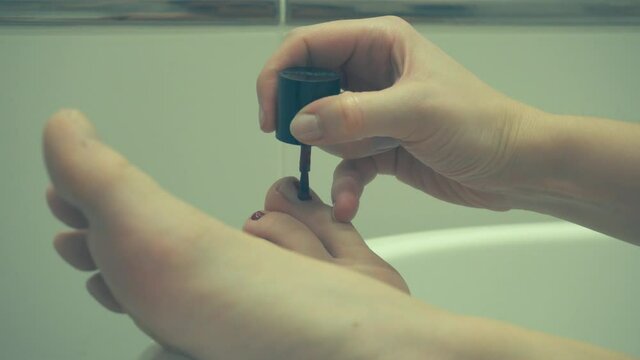 A woman in the bathroom makes herself a pedicure and paints her toe nails with red nail polish.

