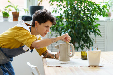 Young female artisan concentrated on modeling jug from raw clay while pottery lesson or workshop in...