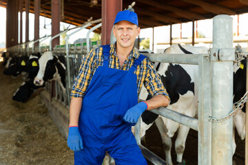 Portrait of positive dairy farm worker in blue overalls against the background of cows in a stall