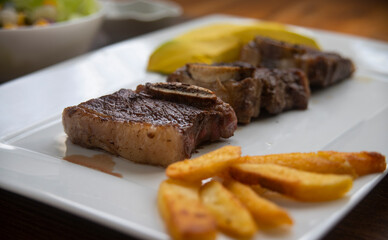 ROASTED BEEF, SERVED ON A WHITE PLATE AND LETTUCE, TOMATO AND CORN SALAD, ACCOMPANIED WITH FRENCH FRIES AND AVOCADO