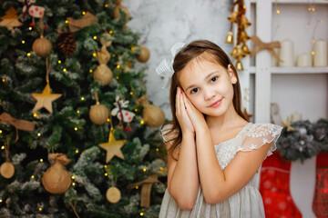 Selective focus. Portrait of a brunette girl in a silver dress against the background of lights of garlands and a Christmas tree decorated for Christmas. Dreamily folded her hands, palms at her face.