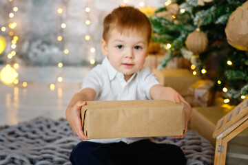 Selective focus. A boy is sitting on the floor with a gift box for a room decorated for Christmas against the background of lights of garlands. He hands the gift to the camera.