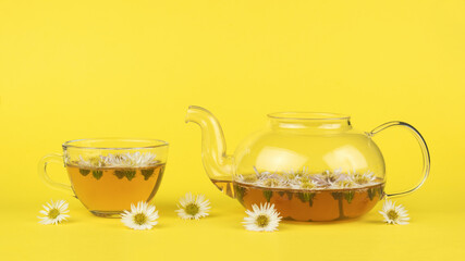 Teapot and glass cup with tea and flowers on a yellow background.