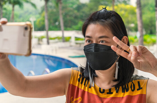 Portrait happy Asian middle-aged woman with black colour face mask taking selfie in a resort while travelling in Thailand, showing two fingers gesture, new normal lifestyle.
