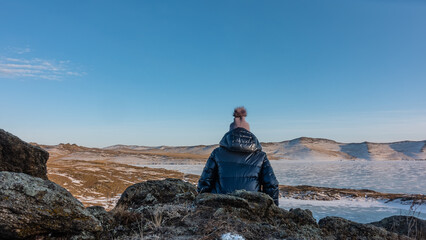 A girl in a down jacket and hat is sitting on a granite rock. View from the back. In front of her is a frozen lake with snow patterns on the ice. A mountain range against a blue sky. Baikal