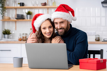 Happy charming family couple in santa hat hugging looking at laptop screen