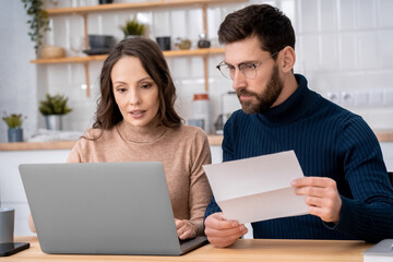 Family married couple looking at computer pc screen making the utility payments sitting at home kitchen. Wife holding paper bills and husband paying online using banking application on laptop indoors