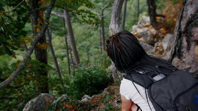 Adventure trip - young woman hiker with dreadlocks sits down on the ground in the forest and looking at the mountainous descent down
