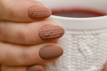 Stylish winter nail design. Knitted fabric texture on women's nails. A woman's hand holds a cup of...