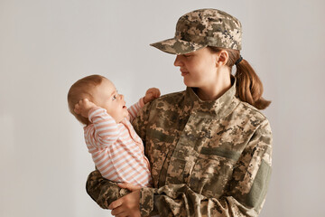 Caucasian female soldier wearing camouflage uniform and cap, standing holding toddler daughter, looking at her baby with love, missing her child while being i army or war.