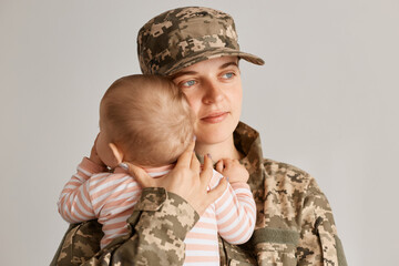 Indoor shot of happy calm woman soldier embracing her little kid in striped sleeper, mother expressing positive emotions, hugging her child with great love.
