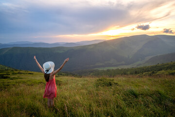 Fototapeta na wymiar Back view of young happy woman traveler in red dress standing on grassy hillside on a windy evening in summer mountains with outstretched arms enjoying view of nature at sunset