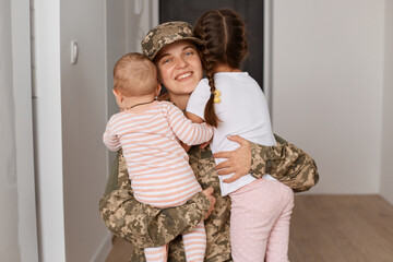 Portrait of positive smiling military mother wearing camouflage uniform and hat, embracing children...