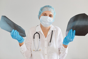 Indoor shot of confused female doctor wearing medical uniform, cap and gloves, holding two x rays, dont know diagnosis, radiologist standing with doubtful expression.