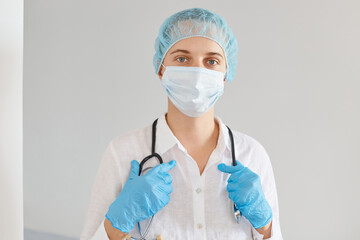 Good looking young adult Caucasian woman doctor wearing medical cap, gloves and protective mask, standing with calm facial expression, waiting her patients.