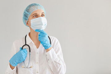 Pensive female doctor wearing medical uniform, cap and surgical mask, standing , holding chin, thinking about new methods of treatment, having thoughtful facial expression.