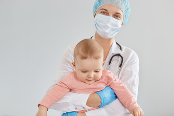 Smiling attractive female doctor pediatrician wearing medical cap, gloves and protective mask, standing with toddler baby girl in hands, routine medical examination by neuropathist.