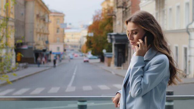 Serious young successful lady business woman girl in blue suit stands on balcony talking mobile phone. Female consultant speaks with smartphone. Buyer online talk cellphone standing on city background