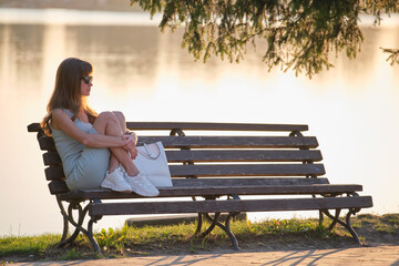 Lonely woman sitting alone on lake shore bench on warm summer evening. Solitude and relaxing in nature concept