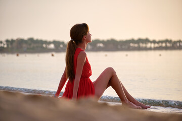 Fototapeta na wymiar Young happy woman in red dress relaxing on sandy beach by seaside enjoying warm tropical morning